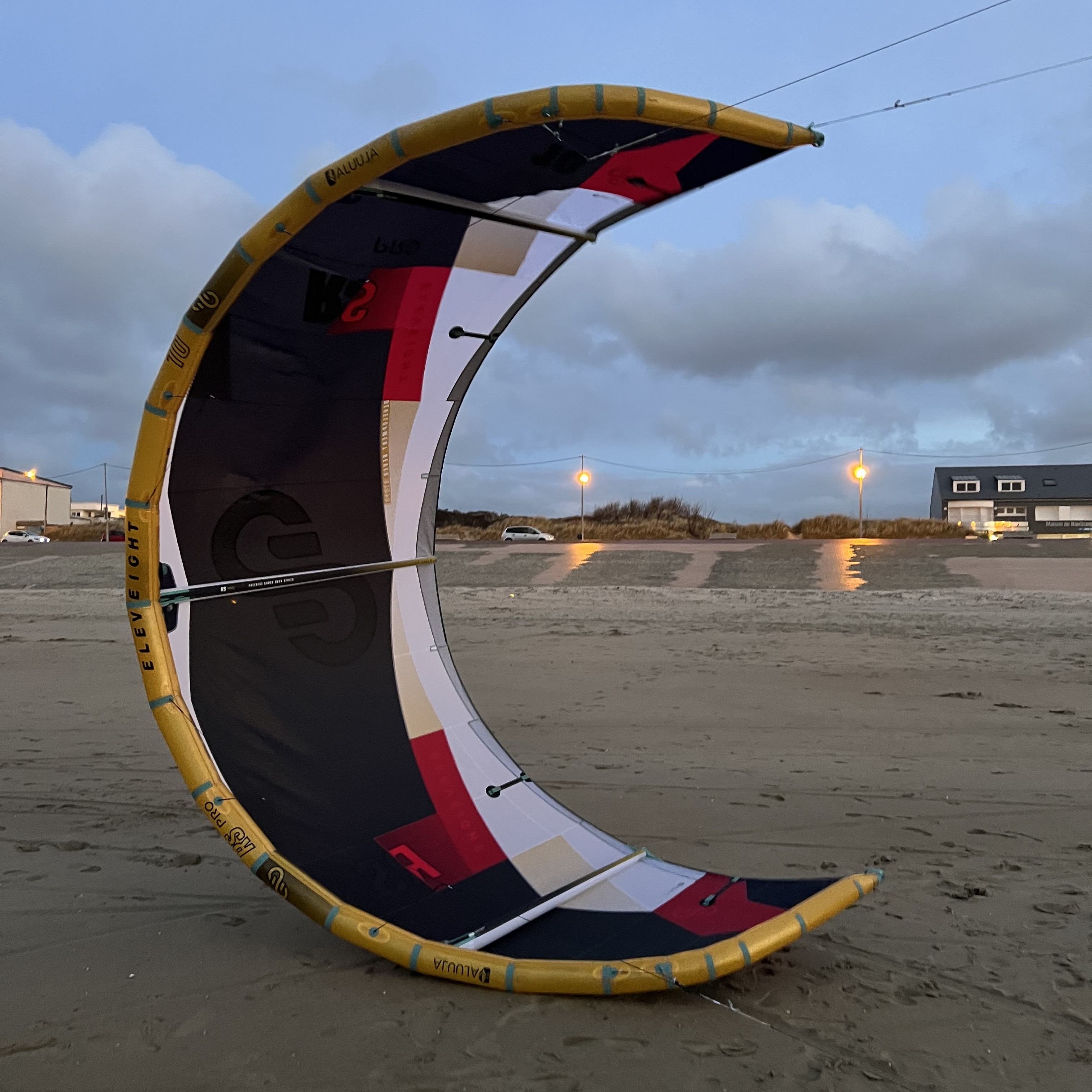Aile de kitesurf Eleveight RS PRO V1 2025 posée verticalement sur le sable au coucher du soleil, avec maisons et lumières urbaines en arrière-plan, idéale pour le freeride et le big air.